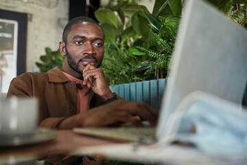 Portrait of modern African American man working with laptop while sitting at table in eco friendly green cafe interior, copy space