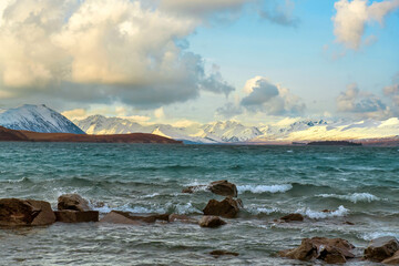 Sticker - Rocks in turquoise water, waves in Lake Tekapo, background is Mount Cook Mountains. On a beautiful cloudy day on the South Island of New Zealand.