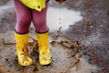 Close-up Of Little Toddler Girl Wearing Yellow Rain Boots And Walking During Sleet On Rainy Cloudy Day. Cute Child In Colorful Clothes Jumping Into Puddle, Splashing With Water, Outdoor Activity