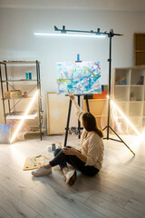 Wall Mural - Female painter. Brush stroke. Creating picture. Art school. Leisure hobby. Thoughtful inspired woman sitting on floor painting abstract artwork on easel in light interior studio.