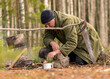 © ANDA - man in the forest trying to light a campfire, a tree branch with a pot over the campfire, blurred forest background, bonfire and smoke, autumn time