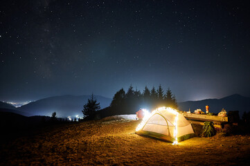 Wall Mural - Fantastic view of night starry sky over mountains with illuminated touristic tent. Back view of hiker sitting near campfire under blue sky with stars. Concept of travelling, hiking and night camping.