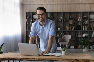 Poster - Portrait of smiling young Caucasian businessman stand at desk in home office work online on laptop gadget. Happy man use computer consult client or customer distant on device. Technology concept.