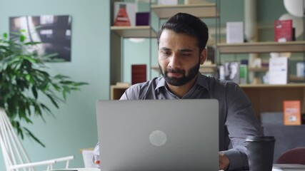 Wall Mural - Young bearded indian businessman working on laptop in modern office lobby space. Indian executive using computer remote studying, watching online webinar, virtual training from homeoffice.