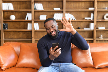 african american man sitting at home on leather sofa talking chatting via video call using app on sm