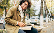 © iuricazac - Horizontal image of a young man reading a book outdoors. College male student learns on campus in the autumn street. Smiling smart guy with curly hair wears spectacles and reading books in the city.