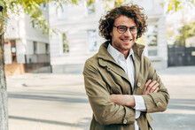 A Young Successful Businessman In Eyewear Smiling Broadly Posing Outdoors. Male Entrepreneur Resting In The City Street. Smart Guy In Casual Wears Spectacles With Curly Hair Walking Outside After Work