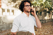 © iuricazac - Outdoor shot of a smart young man talking on mobile phone in the street. Handsome male with curly hair in a white shirt and eyeglasses making a call on his mobile phone in the city street.