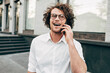 © iuricazac - Businessman with curly hair smiling broadly while talking with his friend on mobile phone outdoor. A cheerful curly male wearing a white shirt has a joyful expression during speaking on a cellphone.