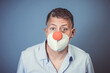 Middle aged man with nose mouth protection and red clown nose and blue shirt in front of blue background