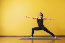 Attractive Woman In Black Sportswear Practicing Yoga Performs Virabhadrasana Exercise, Warrior Pose 2, Against A Yellow Wall With Copy Space