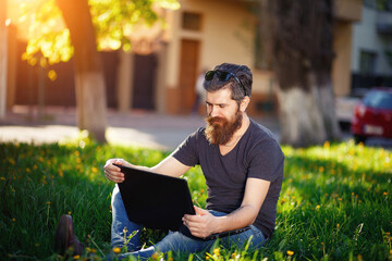 A man with a long beard womanizer and dark hair with gray hair in a shirt and jeans sits on green grass in a park with a laptop and watches funny videos