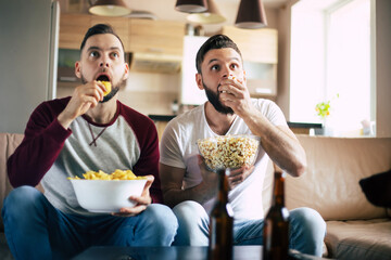 Wall Mural - Two young happy bearded friends watching TV or some sport match while sitting on the couch at home on weekend and drinking beers and eating snacks