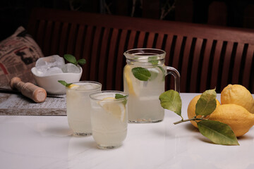 Pitcher and two glasses of ice cold lemonade and lemons on the table. Refreshing mint lemonade in glasses on a white marble table and a wooden bench behind.