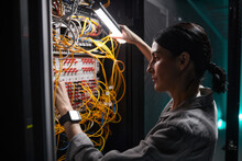 Side View Portrait Of Female Network Engineer Connecting Cables In Server Cabinet While Working With Supercomputer In Data Center