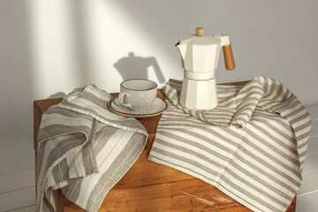 Italian coffee maker with wooden handle and white coffee craft cup on solid wood stump decorated with tea towels in creamy tones on sunlight creating beautiful shadows on the wall