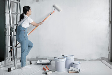 girl paints a white wall with a roller. Repair of the interior. Young female decorator painting a wall in the empty room, concept builder or painter in helmet with paint roller over the empty room