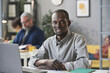 © AnnaStills - Portrait of African young businessman smiling at camera while sitting at the table in front of the laptop at office