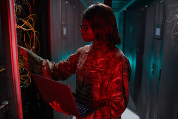 Side view portrait of African-American woman managing computer network while working with supercomputer in server room lit by red light, copy space