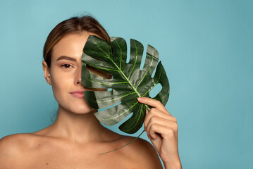 Wall Mural - A lovely female model covers part of her face with a tropical palm leaf. Skin care, hydration. Cosmetics with natural ingredients