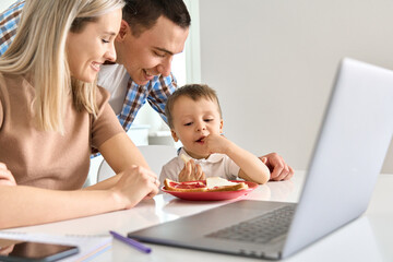 Wall Mural - Happy young family couple with toddler kid son eating toasts with jam using laptop in kitchen. Cute small child boy tasting breakfast snack with parents working from home on computer.