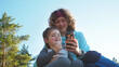 Close up. Happy curly hair mother with little daughter browsing chatting playing serfing network with mobile phone apps during sunset on spring camping vacation. Concept of nature quality family time
