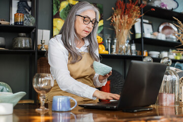 Wall Mural - Beautiful mature lady using modern laptop while receiving new goods for decor shop. Grey-haired saleswoman wearing eyeglasses, white shirt and beaige apron.