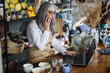 © sofiko14 - Mature saleswoman talking on mobile and typing on laptop while sitting at decor store. Pretty woman accepting order by modern smartphone.