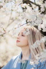 Stylish beautiful woman sensually posing among blooming apple branches in sunny spring, calm moment. Portrait of happy young blonde female in blue jacket embracing in white flowers