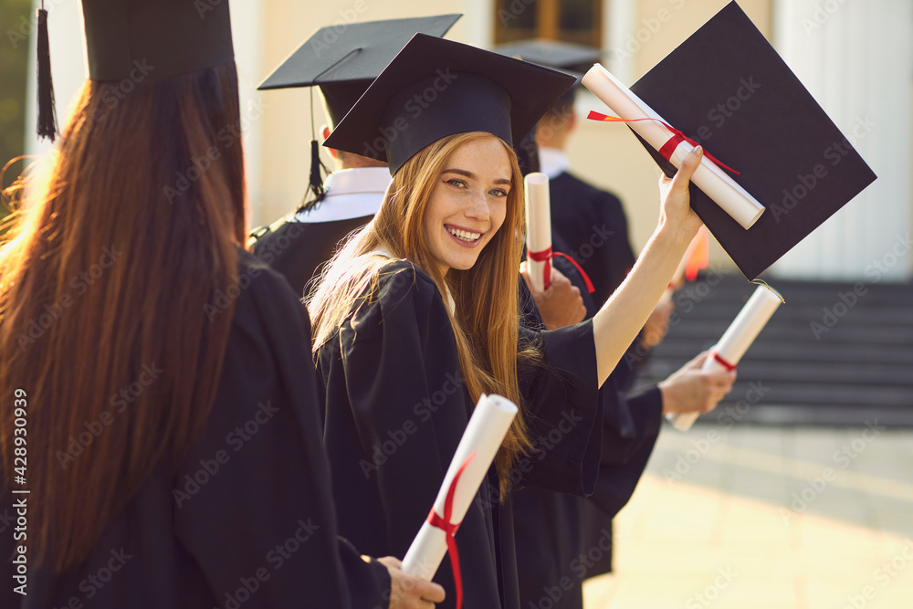 Obraz Successful graduation from university. Smiling beautiful girl university or college graduate standing with diploma and looking at camera over mates around and university at background fototapeta, plakat