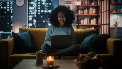 Aufkleber - Beautiful Authentic Latina Female Sitting on Couch Sofa in a Cozy Dark Living Room in the Evening and Using Laptop Computer at Home. She's Browsing the Internet and Checking Videos on Social Networks.