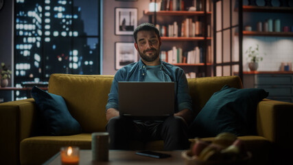 Aufkleber - Handsome Caucasian Man Working on Laptop Computer while Sitting on a Sofa in Dark Cozy Living Room in the Evening. Freelancer Working From Home. Browsing Internet, Using Social Networks, Having Fun.