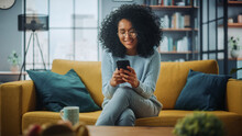 Portrait Of A Beautiful Authentic Latina Female In A Stylish Cozy Living Room Using Smartphone At Home. She's Browsing The Internet And Checking Videos On Social Networks And Having Fun.