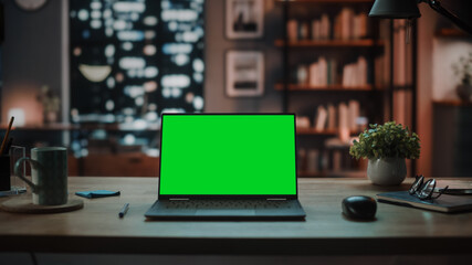 Wall Mural - Laptop Computer with Mock Up Green Screen Display Standing on the Table in Cozy Living Room. In the Background Stylish Modern Home Office Studio in the Evening with Big Cityscape Window.