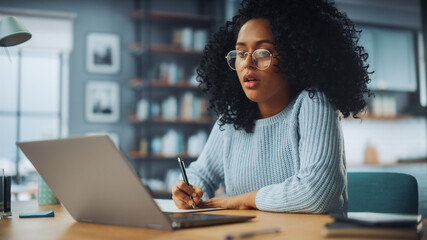 Beautiful Authentic Latina Female Sitting at a Desk in a Cozy Living Room and Following a Video School Lesson on Laptop Computer. Student Studying Remotely. Chat with Friends on Social Network.