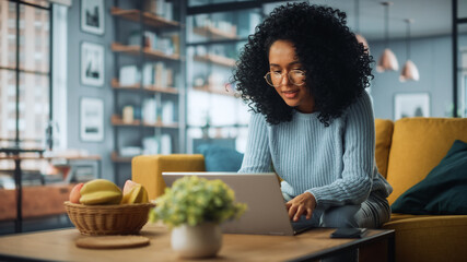 Aufkleber - Beautiful Authentic Latina Female Sitting on a Sofa in a Cozy Living Room and Using Laptop Computer at Home. Happy Freelancer Female Browsing Internet and Social Networks.
