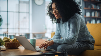 Aufkleber - Beautiful Authentic Latina Female Sitting on a Sofa in a Cozy Living Room and Using Laptop Computer at Home. Happy Freelancer Female Browsing Internet and Social Networks.