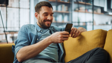 Aufkleber - Happy Handsome Caucasian Man Using Smartphone in Cozy Living Room at Home. Man Resting on Comfortable Sofa. He's Browsing the Internet and Checking Videos on Social Networks and Having Fun.