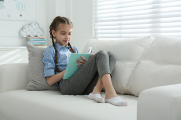 Cute little girl reading book on sofa at home