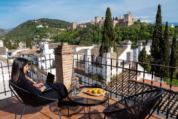 Female travel blogger working on laptop while sitting on terrace with view of Alhambra fortress in Granada, Spain