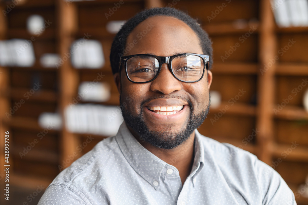 Close-up portrait of a happy African-American young man with friendly wide toothy smile, a mixed-race bearded guy wearing stylish eyeglasse and shirt looks into camera, employee profile photo - obrazy, fototapety, plakaty 