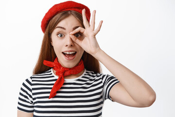 Cheerful young woman in french red beret and striped t-shirt, look through okay sign, make OK gesture over eye and gasping excited, white background