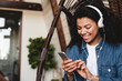 © InsideCreativeHouse - Excited afro young woman listening to the music on her phone in living room. Happy relaxed african woman wear wireless headphones listening to lounge music relaxing after work