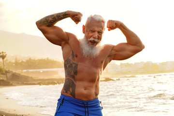 happy fit senior man showing his muscle on the beach after outdoor workout, during sunset time.