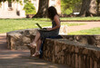A female college student sits in the shade while working on a laptop computer. 