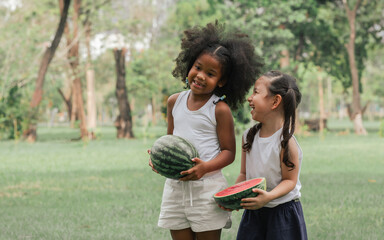 Two African and Caucasian white little girls smiling, laughing with happiness together, carrying watermelon while playing and standing in outdoor green park. Education, Diversity, Fruits Concept.