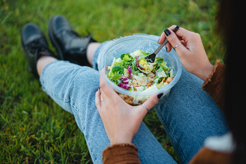 Wall Mural - Close-up of a salad bowl on teenager girl legs. She is sit on grass enjoying nature. Horizontal shot.