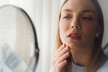 Young beautiful woman looking herself in the mirror at home. Worried about acne caused by wearing a mask. Maskne. Problems with acne.