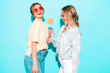 Wall Mural - Two young beautiful blond smiling hipster female in trendy summer clothes. Sexy carefree women posing near blue wall in studio. Trendy and positive models having fun with candy sweet lollipop