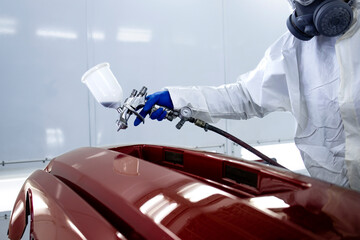 Car painter in protective clothes and mask painting automobile bumper with metallic paint and varnish in chamber workshop.